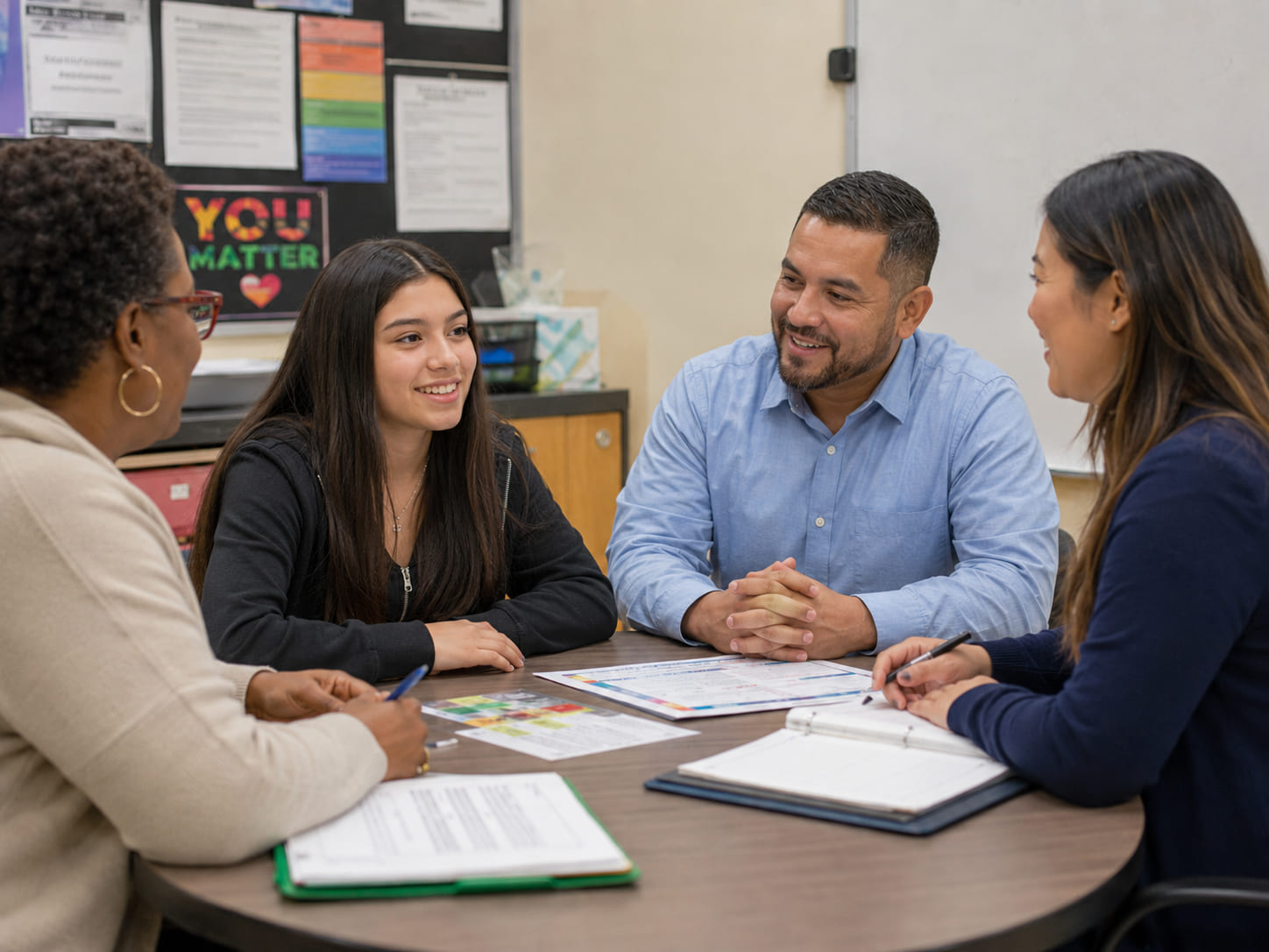 A group sits around a table during an IEP meeting in a school conference room. A high school female student smiles while participating in the conversation with a parent and school staff, with papers, folders, and binders spread across the table.